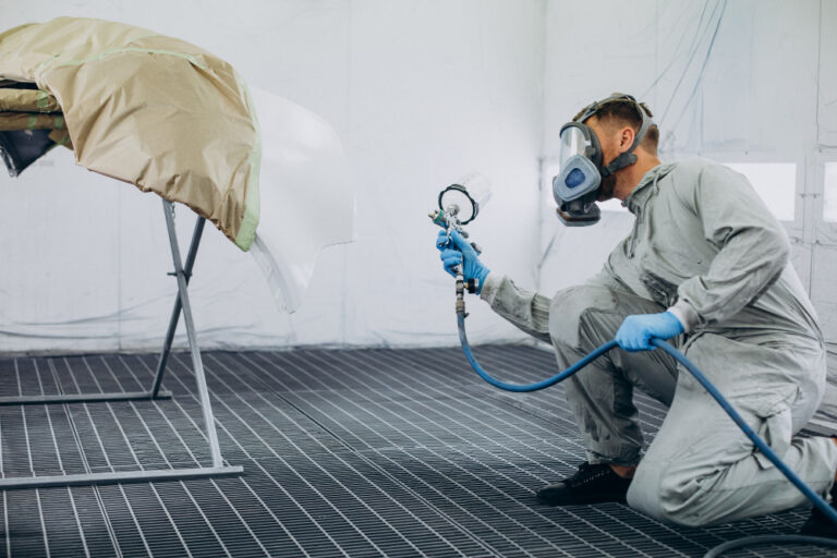 Sistema de exaustão para cabine de pintura: homem usando roupa de proteção e máscara, pintando peça automotiva.