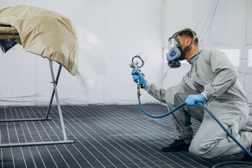 Sistema de exaustão para cabine de pintura: homem usando roupa de proteção e máscara, pintando peça automotiva.